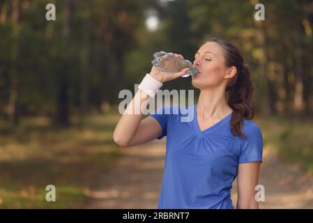 Die schlanke junge Frau trägt Sportbekleidung, die Wasser in Flaschen trinkt, während sie auf einer Waldstrecke pausiert, um sich während eines Trainings auf dem Land zu rehydrieren Stockfoto