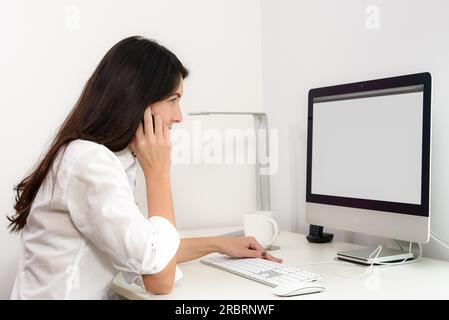 Eine Geschäftsfrau, die in ihrem Büro arbeitet, sitzt an einem Schreibtisch vor ihrem Desktop-Computer und hat einen leeren Bildschirm, der für den Zuschauer sichtbar ist, während sie über sie chattet Stockfoto