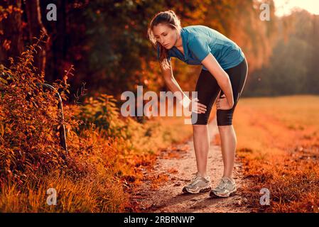 Junge Sportlerin, die eine Pause vom Training im Stehen macht und ihre Hände auf den Knien auf einer ländlichen Strecke durch üppiges Ackerland in einem gesunden und Stockfoto