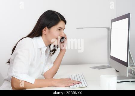 Eine Geschäftsfrau, die in ihrem Büro arbeitet, sitzt an einem Schreibtisch vor ihrem Desktop-Computer und hat einen leeren Bildschirm, der für den Zuschauer sichtbar ist, während sie über sie chattet Stockfoto