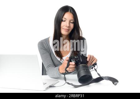 Attraktive junge Fotografin, die ein Bild auf der Rückseite ihrer Kamera abliest, während sie mit der Kamera an ihrem Schreibtisch vor ihrem Laptop sitzt Stockfoto