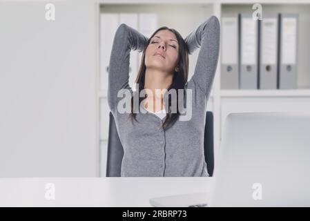Junge Geschäftsfrau genießt einen entspannten Moment, sitzt in ihrem Stuhl an ihrem Schreibtisch mit den Händen hinter dem Kopf und verschlossenen Augen Stockfoto