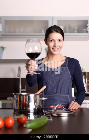 Lächelnde junge Hausfrau feiert mit Rotwein, der ihr Glas erhebt, um auf die Kamera anzustoßen, während sie in der Küche am Herd steht und das Abendessen zubereitet Stockfoto