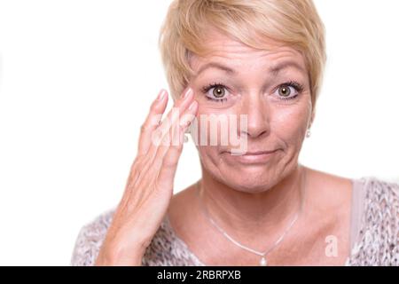 Attraktive ältere blonde Frau mit einem breiten Gesichtsausdruck und ihrer Brille auf dem Kopf, die ihren Teint prüft, während sie in den Spiegel schaut Stockfoto