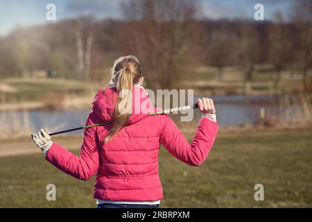 Rückansicht einer blonden Golferin in dunkelrosa Jacke, die einen Golfschläger auf der Schulter hält und aus der Ferne blickt, in Half Body Shot aufgenommen Stockfoto