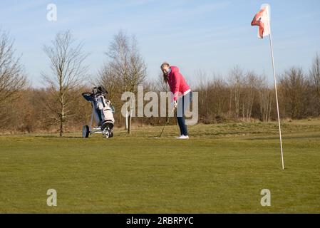 Junge Golferin in einem Golfsporttraining, trägt Jacke und Jeans, schlägt Golfball auf dem Cup mit Flagge Stockfoto