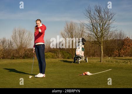 Junge Golferin in Jacke und Jeans mit Putter und Daumen nach oben mit Golfwagen an der Seite Stockfoto