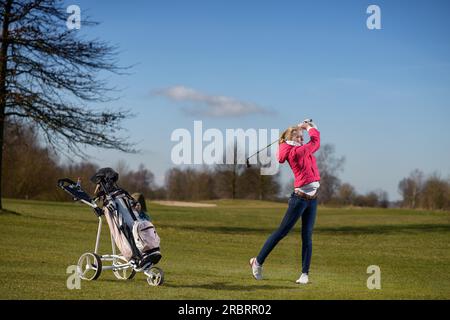 Attraktive, schlanke junge Golferin, die einen Annäherungsschuss auf dem Fairway spielt und den Flug ihres Balls nach dem Schlaganfall mit ihrer Golftasche beobachtet Stockfoto