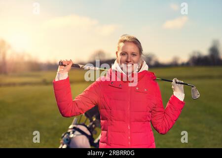 Nahaufnahme lächelnde junge Golferin in roter Jacke mit einem Golfschläger über der Schulter, während sie in die Kamera schaut Stockfoto