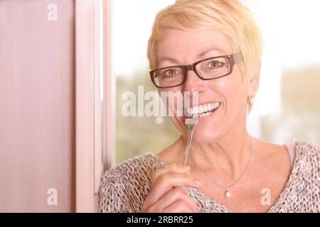 Nahaufnahme Happy Middle Age Blond Woman, trägt eine Brille, beißt in die Gabel, während er in die Kamera schaut Stockfoto