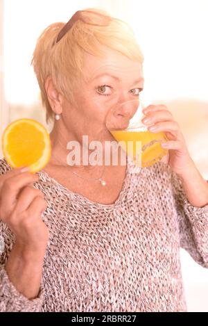 Nahaufnahme einer mittelalterlichen blonden Frau, die Orangensaft trinkt und ein Orangenobststück in der Hand hält, während sie sich an die Wand lehnt und in die Kamera schaut Stockfoto