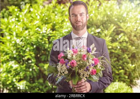 Hübscher, bärtiger junger Mann in einem Anzug mit einem Strauß frischer Blumen, vielleicht ein Verehrer oder Verehrer, der zu einem Date, Valentinstag, einem Jahrestag anruft Stockfoto
