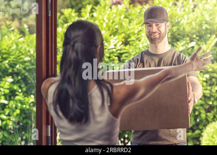 Eine Frau nimmt die Lieferung eines großen Pakets entgegen und hält ihre Arme an den lächelnden freundlichen Zusteller, während er es übergibt, Blick von hinten über sie Stockfoto