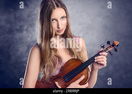 Attraktive junge Musikerin, die ihre Geige in ihrem Arm hielt, während sie mit einem Lächeln auf blauem Hintergrund in die Kamera schaute Stockfoto