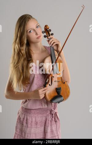 Attraktive junge Musikerin, die ihre Geige in ihrem Arm hielt, während sie mit einem Lächeln in die Kamera blickte, über einem grauen Hintergrund Stockfoto