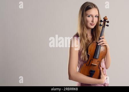 Attraktive junge Musikerin, die ihre Geige in ihrem Arm hielt, während sie mit einem Lächeln in die Kamera blickte, über einem grauen Hintergrund Stockfoto