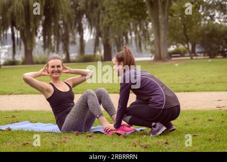 Fröhliche Athletikerin lächelt in die Kamera, während sie mit Hilfe ihrer Freundin im Park ein Curl-Up-Training macht Stockfoto
