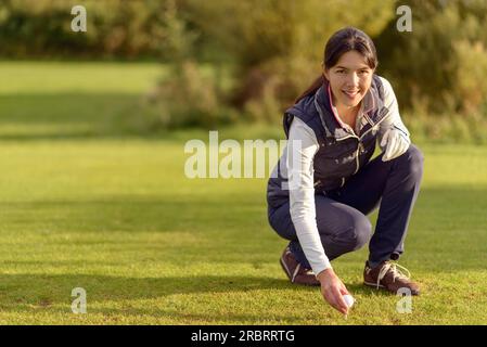Lächelnde junge Golferin bückt sich auf dem Fairway und legt einen Ball auf ein T-Shirt auf einem üppig grünen Golfplatz, während sie sich auf ein Loch vorbereitet Stockfoto