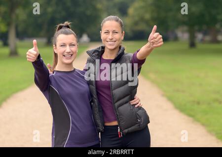 Halbkörperaufnahme von zwei glücklichen, gesunden Frauen im Park, die nach ihrem Training Daumen nach oben in die Kamera zeigen Stockfoto