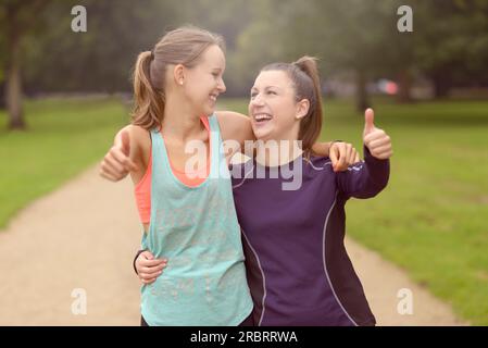 Halbkörperaufnahme von zwei glücklichen, gesunden Frauen im Park, die nach ihrem Training Daumen nach oben in die Kamera zeigen Stockfoto