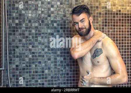 Freundlicher junger Mann mit Vollbart, der sich in der Dusche einseift und an die Kamera lächelt, Oberkörper mit Kopierbereich Stockfoto