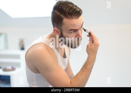 Junger Mann rasieren über seinen Bart mit einem manuellen Rasiermesser spiegelt sich in den Spiegel im Bad in einem Pflege und Hygiene-Konzept Stockfoto