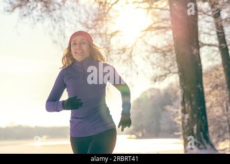 Ganzkörperaufnahme einer hübschen Athletikerin, die im Winter mit Kopfhörern und Winterkleidung joggt, die Kamera anlächelt, mit Kopierbereich Stockfoto