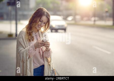 Attraktive junge Frau, die im Morgenlicht am Rande einer urbanen Straße steht und mit einem fröhlichen Lächeln eine Nachricht auf ihrem Mobiltelefon abliest Stockfoto