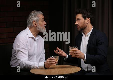 Männer mit Whiskey-Gläsern, die sich drinnen an einem Holztisch unterhalten Stockfoto