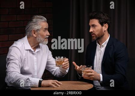 Männer mit Whiskey-Gläsern, die sich drinnen an einem Holztisch unterhalten Stockfoto