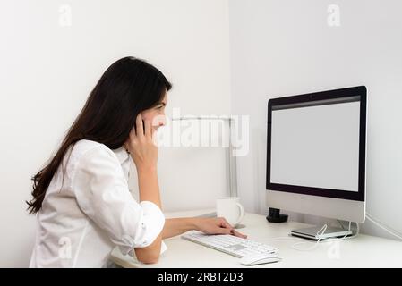 Eine Geschäftsfrau, die in ihrem Büro arbeitet, sitzt an einem Schreibtisch vor ihrem Desktop-Computer und hat einen leeren Bildschirm, der für den Zuschauer sichtbar ist, während sie über sie chattet Stockfoto