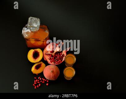 Zwei Gläser und eine Karaffe aus Glas mit hausgemachtem Likör auf schwarzem Hintergrund, neben Stücken eines reifen Pfirsichs und eines zerbrochenen Granatapfelapfel. Draufsicht, flach Stockfoto