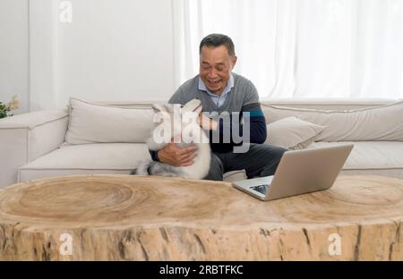 Großvater im Ruhestand verbrachte Zeit mit seinem Haustier im Wohnzimmer zu Hause. Laptop-Computer sind auf einem Tisch aus einem großen Baumstamm. Stockfoto