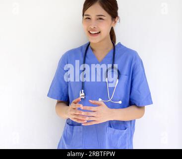 Junger asiatischer Physiotherapeut in blauer Uniform und Stethoskop stehen lächelnd vor der weißen Wand. Stockfoto