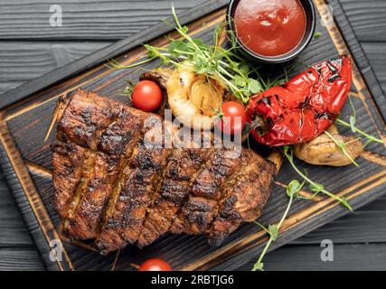 Gegrillte Schweinerippchen mit Kirschtomaten, Pfeffer und Barbecue-Sauce auf Holzbrettern vor dunklem Hintergrund. Draufsicht, flach liegend Stockfoto