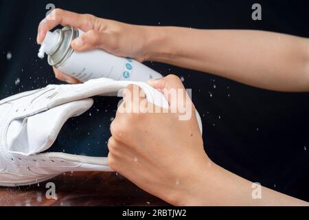 Eine Frau sprüht Reinigungsschaum auf weiße Lederschuhe. Pflege von Lederschuhen. Stockfoto