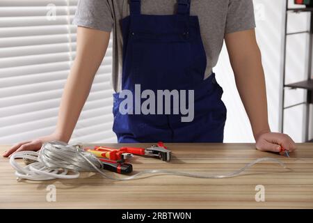 Professioneller Elektriker in Uniform, Werkzeug und Draht auf Holztisch in Innenräumen, Nahaufnahme Stockfoto