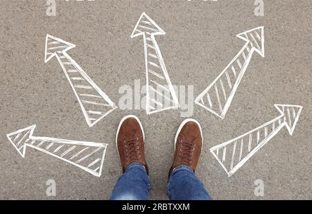 Den Beruf der Zukunft wählen. Mann steht vor gezeichneten Schildern auf Asphalt, Draufsicht. Pfeile, die in verschiedene Richtungen zeigen und Diversität symbolisieren o Stockfoto