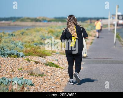 Rückansicht einer jungen Frau mit Kopfhörern, Trainingsanzug und Schultertasche, die im Sommer in Großbritannien entlang einer Straße am Meer läuft. Stockfoto