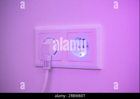 Blau und violett beleuchtete Buchse mit eingestecktem Stecker zur Darstellung des Stromverbrauchs und der elektrischen Energie aus einer Steckdose Stockfoto