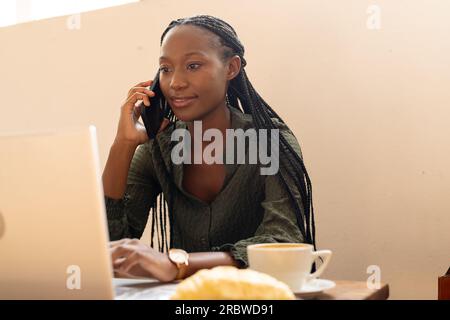 Eine afroamerikanische Frau, die ein Notebook benutzt und in einem Café mit einem Smartphone spricht Stockfoto
