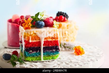 Verschiedene Scheiben Kuchen auf einem weißen Tablett: regenbogenkuchen, Himbeerkuchen und Mandelkuchen. Süßigkeiten dekoriert mit frischen Beeren und Blumen für den Urlaub Stockfoto