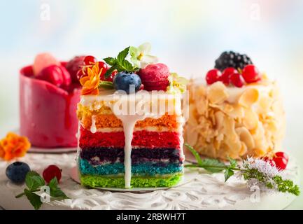 Verschiedene Scheiben Kuchen auf einem weißen Tablett: regenbogenkuchen, Himbeerkuchen und Mandelkuchen. Süßigkeiten dekoriert mit frischen Beeren und Blumen für den Urlaub Stockfoto