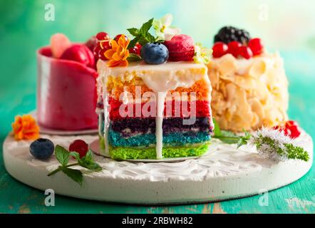 Verschiedene Scheiben Kuchen auf einem weißen Tablett: regenbogenkuchen, Himbeerkuchen und Mandelkuchen. Süßigkeiten dekoriert mit frischen Beeren und Blumen für den Urlaub Stockfoto