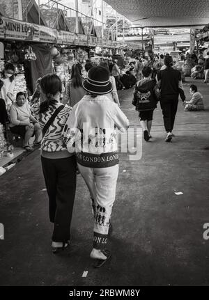 Zwei Frauen, eine in chinesischer Kleidung, die London darstellt, gehen Arm in Arm an Verkaufsständen in Con Market, Danang, Vietnam vorbei. Stockfoto