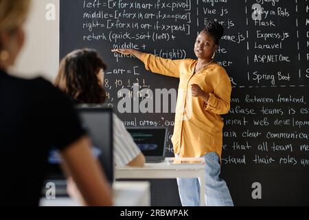 Afroamerikanischer Lehrer, der mit Formeln auf die Tafel zeigt und den Schülern im Unterricht Material erklärt Stockfoto
