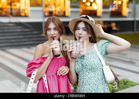 Zwei junge, schön lächelnde Frauen in trendigen Sommerkleidung. Unbeschwerte Frauen posieren im Freien. Positive Modelle halten und trinken kühlen Drink in Kunststoff Stockfoto