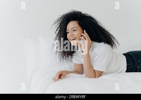Junge Frau unterhält sich mit ihrem Freund, morgens im Bett, lacht über Witze, lässige Kleidung, Wohngegend an der weißen Wand. Kommunikationsschwingungen. Stockfoto