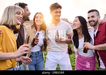 Multiethnische Freunde, die sich bei einer Flasche Bier im Freien unterhalten. Gruppe junger, vielfältiger stehender Menschen. Stockfoto