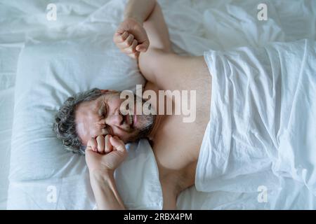 Männlicher Kopf auf Kissen. Ein Mann mit grauen Haaren wacht im Bett auf. Ein älterer Mann wacht im Schlafzimmer auf. Ein erwachsener Mann wacht morgens zu Hause auf. Schlaf gut. Millennials Stockfoto